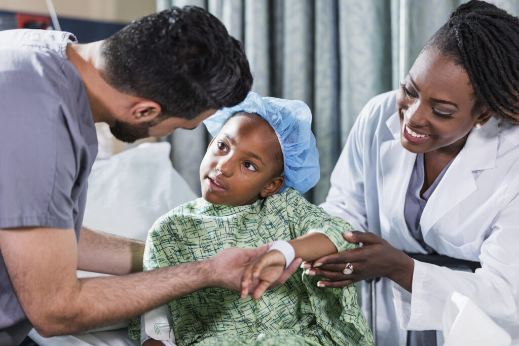 Uma menina afro-americana de 6 anos de idade sentada em uma maca, com um médico e uma enfermeira cuidando dela. Ela está usando uma bata de hospital e uma touca cirúrgica. Uma enfermeira de avental, um jovem do Oriente Médio na faixa dos 20 anos, está de pé ao lado dela, e uma médica, uma mulher afro-americana madura na casa dos 40 anos, está de pé do outro lado.