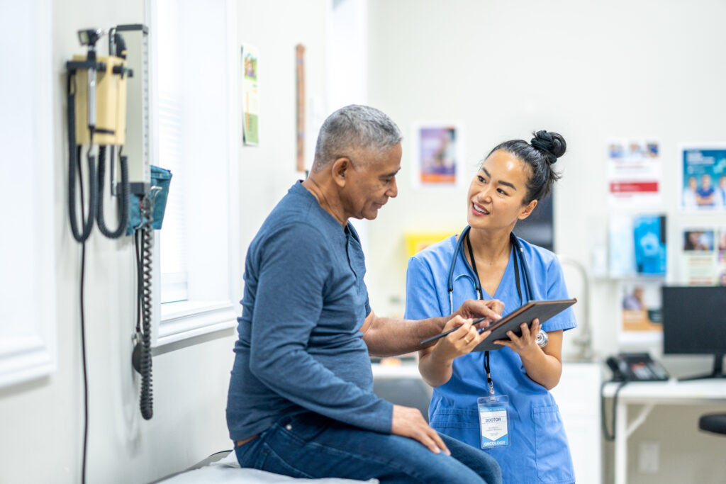 Um senhor idoso de descendência hispânica senta-se em uma mesa de exame durante um check-up de rotina com os médicos. Uma enfermeira, que está vestindo um avental azul, está em pé na frente do senhor enquanto analisa alguns resultados de exames recentes com o paciente em um tablet.
